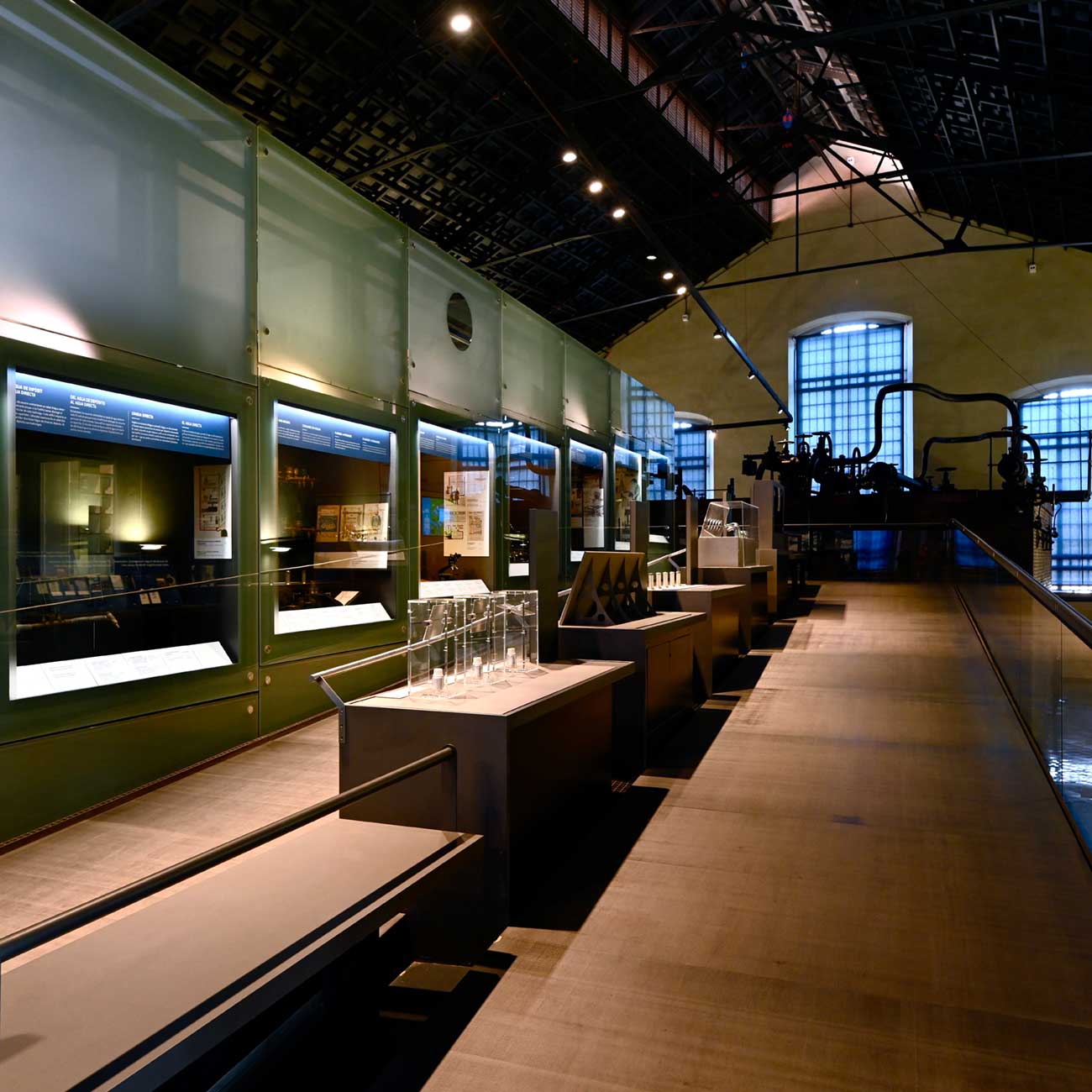 Interior of the exhibition hall, with glass display cases along one side and the original steam machinery preserved at the far end beneath the arched windows.