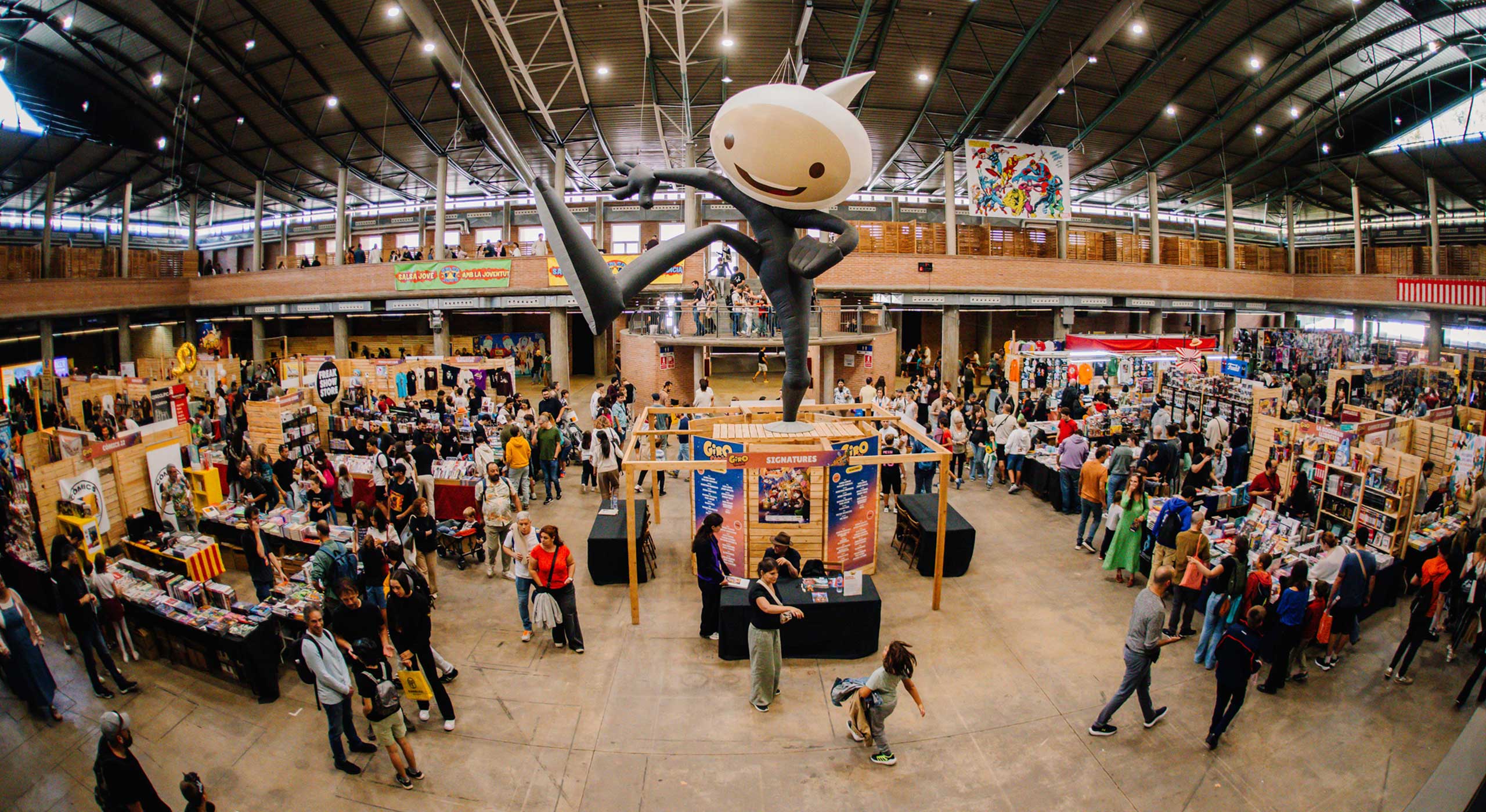 Interior of the Girocòmic fair hall in Girona, with the event's giant mascot sculpture towering over the artist-signing area and rows of comic and manga stalls.
